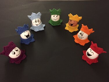 Bunte, gehäkelte Eierwärmer mit aufgemalten Gesichtern auf einer dunklen Oberfläche arrangiert.