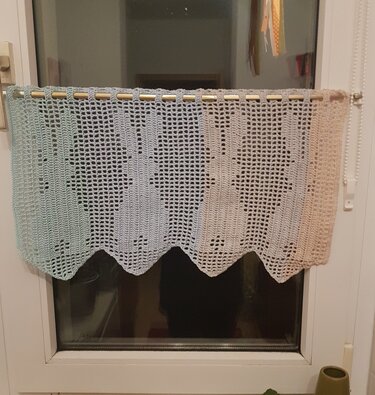 Hell gehäkelte Gardine mit Hasenmotiven im Filetmuster und gezackter Unterkante, an einer Stange vor dem Fenster