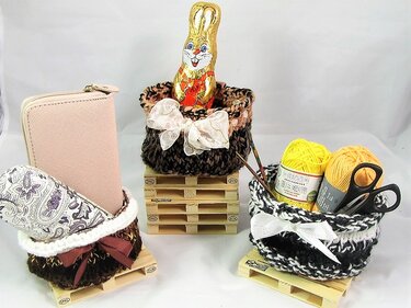 Drei gestrickte Körbchen auf Mini‑Holzpaletten, eines mit Schokoladenhase, die anderen mit Garn und Portemonnaie