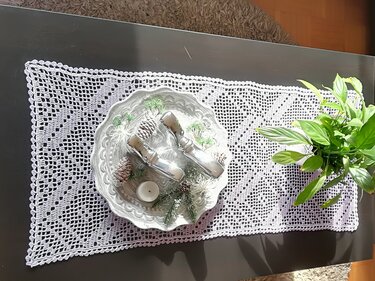 Weißer gehäkelter Tischläufer im Filetmuster mit Diamantmotiven auf dunklem Tisch, dekoriert mit Schale und Pflanze