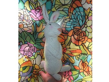Weißer gehäkelter Amigurumi-Hase mit rosa Nase und Ohren, stehend, vor buntem Blumenmuster, Hand stützt ihn unten