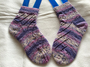 Handgestricktes Paar Damensocken in Violett, Rosa und Beige mit diagonalem Lochmuster auf hellem Untergrund; blaue Sockenspanner sichtbar.