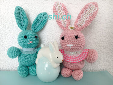 Zwei gehäkelte Amigurumi-Hasen, mintgrün und rosa, mit Perlendetails neben einer weißen Hasenfigur auf einem gepunkteten Ei.