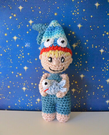 Gehäkelte Amigurumi-Puppe: Junge mit blonden Haaren, blauer Fischmütze und blauem Overall, der einen kleinen gehäkelten Fisch hält, vor sternenblauem Hintergrund.