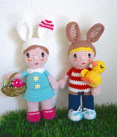 Zwei Amigurumi-Osterhasen, eine mit Eierkorb, die andere mit Küken, auf grünem Untergrund
