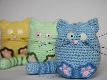 Drei gehäkelte Amigurumi‑Katzen in Blau, Hellgrün und Gelb nebeneinander