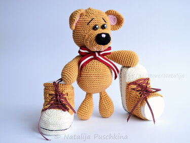 Gehäkelter Teddybär mit rot-weißem Band zwischen zwei gehäkelten Baby-Sneakern in Braun und Weiß.