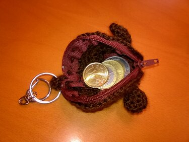 Braune, gehäkelte Münztasche in Schildkrötenform mit rotem Reißverschluss, Schlüsselring und sichtbaren Münzen auf orangefarbenem Untergrund.
