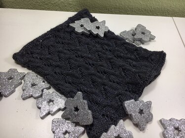 Dunkelgrauer gestrickter Kragenschal mit Zopf- und Lochmuster, dekoriert mit silbernen Sternen auf weißer Unterlage.