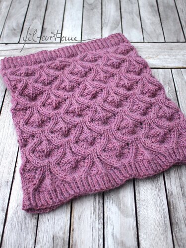 Dusty rose knitted cowl with a raised textured stitch and ribbed edges on a light wooden background.