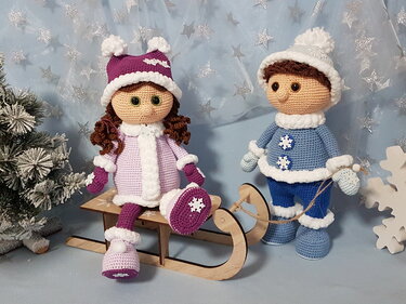 Zwei gehäkelte Amigurumi-Puppen in Winterkleidung, eine sitzt auf einem Holzschlitten