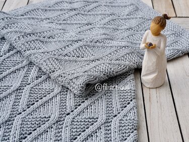 Light gray knitted baby blanket with diamond stitch pattern folded on wooden boards with a small carved wooden figure