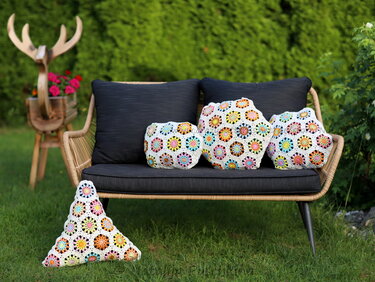 Drei bunte gehäkelte Granny‑Square‑Kissen auf einer dunkelgrauen Gartenbank; ein dreieckiges Kissen liegt im Vordergrund auf dem Rasen