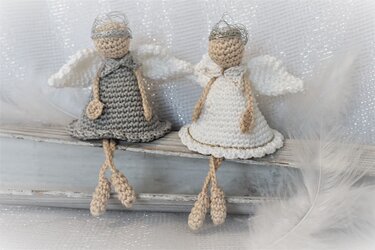 Zwei gehäkelte Amigurumi-Engelchen, eines grau und eines weiß, mit Flügeln und silbernem Kranz, sitzend auf einem Holzbrett