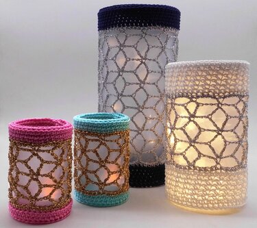 Vier gehäkelte Windlichter in verschiedenen Größen auf hellem Hintergrund, innen beleuchtet. Farben: Rosa, Türkis, Dunkelblau und Weiß mit glänzenden Garnakzenten.