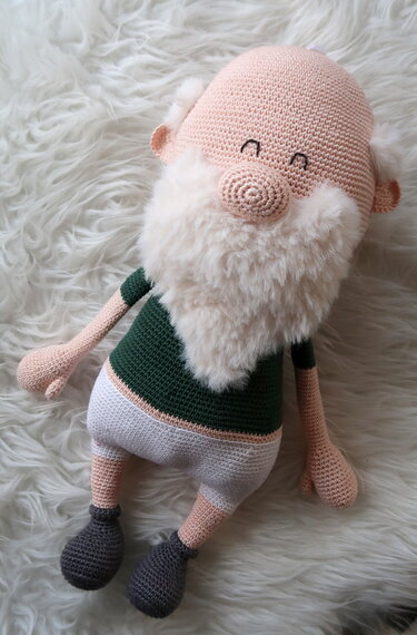 Gehäkelter Wichtel mit weißem, flauschigem Bart, grünem Oberteil und weißer Hose auf weißem Fell.