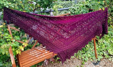 Großes gehäkeltes Dreieckstuch mit offenem Muster und Borte, in Purpurtönen über einer Bank ausgebreitet