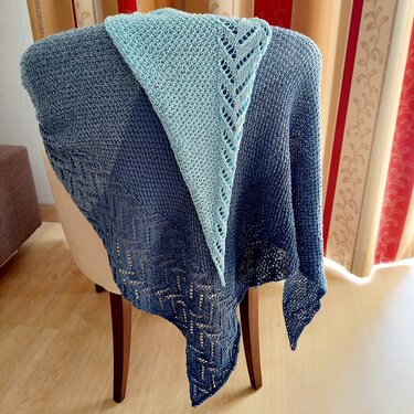 Two-tone blue ombre knitted triangle shawl with eyelet lace border and textured stitches draped over a chair.