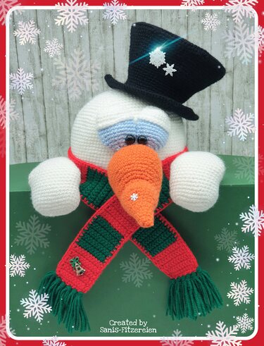 Gehäkelter Schneemannkopf mit schwarzem Zylinder, orangefarbener Nase und rot-grünem Schal vor winterlicher Deko