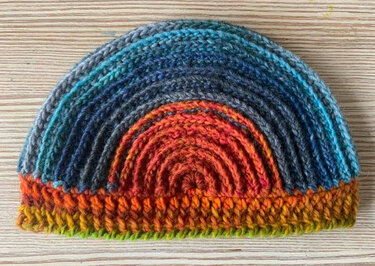 Halbrunde gehäkelte Damenmütze mit Rippenstruktur und Farbverlauf von Orange zu Blau auf hellem Holz