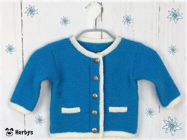 Gestrickte blaue Babyjacke mit weißen Kanten, Knopfleiste und Ziertaschen an einem Kleiderbügel vor hellem Holz mit Edelweiß-Motiven.