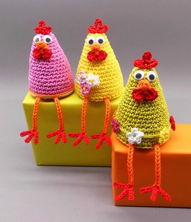 Three pastel crochet chick decorations in pink, yellow and green sitting on yellow and orange boxes