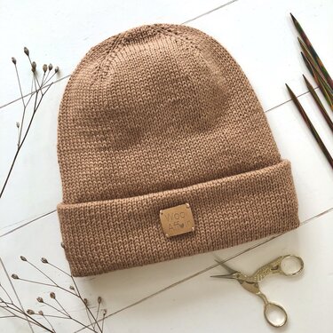 Light brown knitted beanie with a folded ribbed brim, small tag, knitting needles, and scissors on a white wooden background.