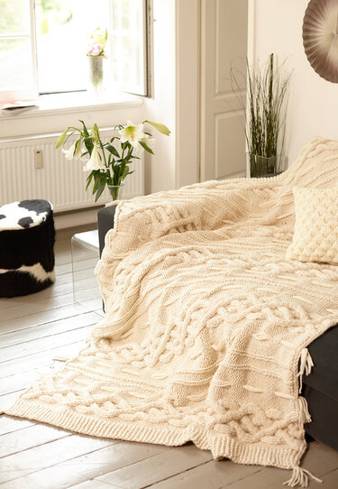 Cremefarbene Strickdecke mit Zopfmuster und Fransen auf einem Sofa