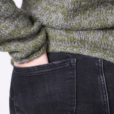 Close-up of a woman wearing a green and grey knitted sweater with a hand in a black jeans pocket.