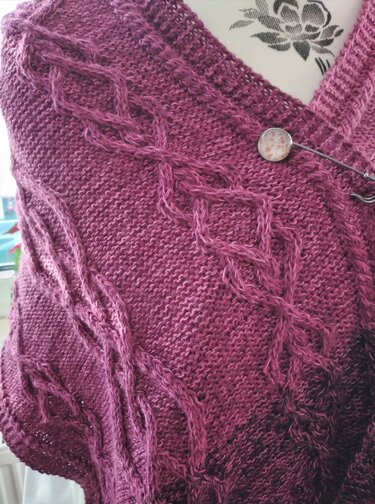 Nahaufnahme eines gestrickten Schals in Beerentönen mit Zopf- und Rautenmuster und einer dekorativen Brosche