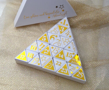 Weißer dreieckiger Adventskalender mit goldenen Zahlen und Sternen auf 25 kleinen Boxen, angeordnet zu einem großen Dreieck auf goldenem Netzstoff.