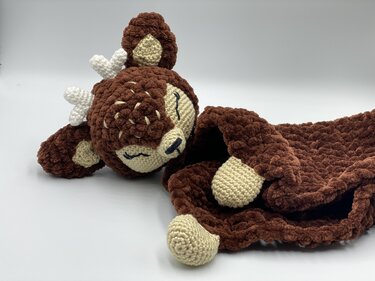 Brown crochet deer snuggle blanket with small white antlers and beige face details lying on a white surface.