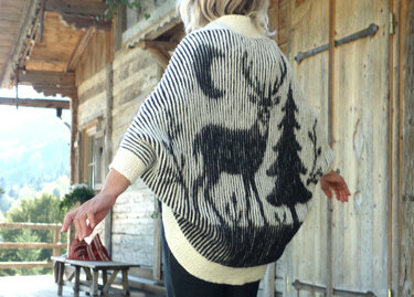 Rückansicht einer Person, die einen schwarz-weißen gestrickten Seelenwärmer mit Hirsch- und Tannenmotiv trägt.