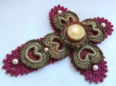 Gehäkeltes Kreuz in Magenta und Goldbraun mit leuchtender LED-Kerze und Perlen auf hellem Hintergrund