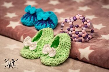 Hellgrüne gehäkelte Babyschuhe mit weißen Schleifen, im Hintergrund türkise Babyschuhe und ein Perlenbeißring auf sternenflauschigem Stoff