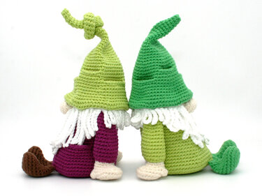 Two crocheted gnomes seen from behind wearing different green hats and white beards, seated on a white background.