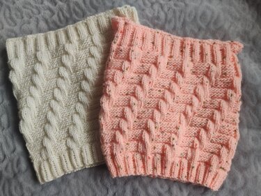 Zwei gestrickte Loopschals in Creme und lachsrosa mit Zopfmuster auf grauem Untergrund