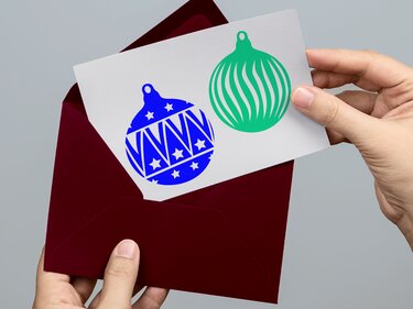 Karte mit zwei Plottmotiven: blaues Weihnachtskugel-Design mit Sternen und Zickzack und grünes Kugel-Design mit geschwungenen Streifen, Hand hält Karte über dunkelrotem Umschlag.