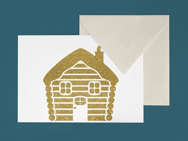 Weiße Grußkarte mit goldglitzernder Blockhütte und beigefarbenem Umschlag auf blaugrünem Hintergrund.