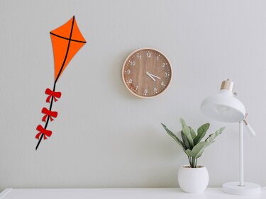 Oranger Drachen mit schwarzen Linien und roten Schleifen am Schwanz an heller Wand neben Uhr und Pflanze