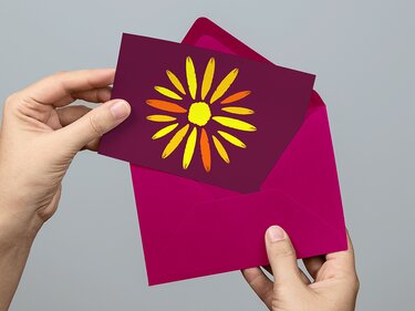Hände halten eine Karte mit gelb-orangem sonnenförmigem Blumenmotiv über einem pinken Umschlag