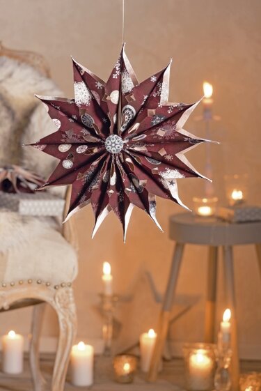Dunkelroter, gefalteter Papierstern mit weißen und silbernen Wintermotiven, hängend vor warmem Kerzenlicht.