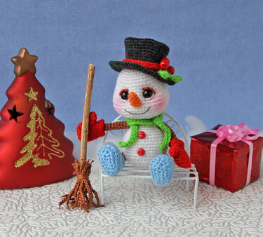 Gehäkelter Amigurumi‑Schneemann mit Zylinder, grünem Schal und Besen neben Geschenk