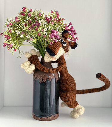 Nahaufnahme eines braunen gehäkelten Amigurumi mit langen Armen und gestreiftem Schwanz neben einer Vase