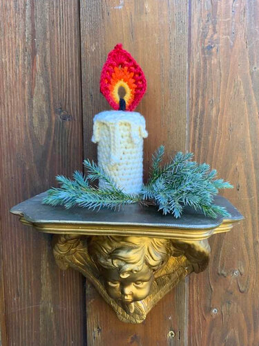 Gehäkelte Kerze mit bunter Flamme auf goldener Wandkonsole mit Tannenzweigen