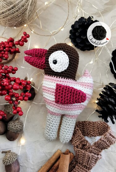 Gehäkelter Amigurumi-Vogel mit braunem Kopf, rosa-weiß gestreiftem Körper, rosa Schnabel und grauen Beinen, dekoriert mit roten Beeren und Zapfen.