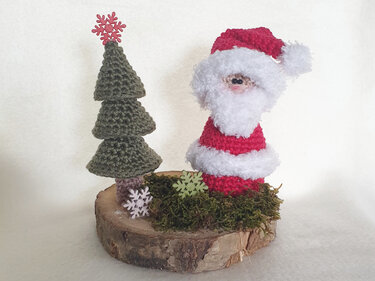 Gehäkelter Weihnachtsmann mit wei
em Bart und roter Mütze neben einem grünen, gestuften Tannenbaum auf einer Holzscheibe mit Moos und Schneeflocken-Dekoration.