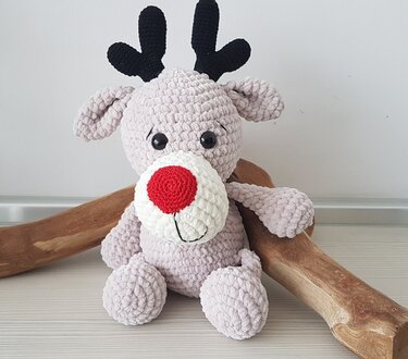 Gehäkeltes Amigurumi-Rentier mit roter Nase und schwarzem Geweih, sitzend auf einem hellen Holztisch neben Dekoholz.