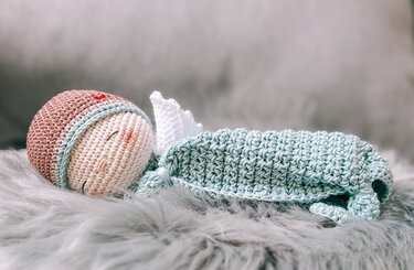 Crocheted sleeping amigurumi baby with brown beanie, blue wrap and white angel wings on grey faux fur.