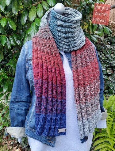 Gehäkelter Schal in Blau, Rot und Grau an einer Schaufensterpuppe mit Jeansjacke vor grünem Blattwerk